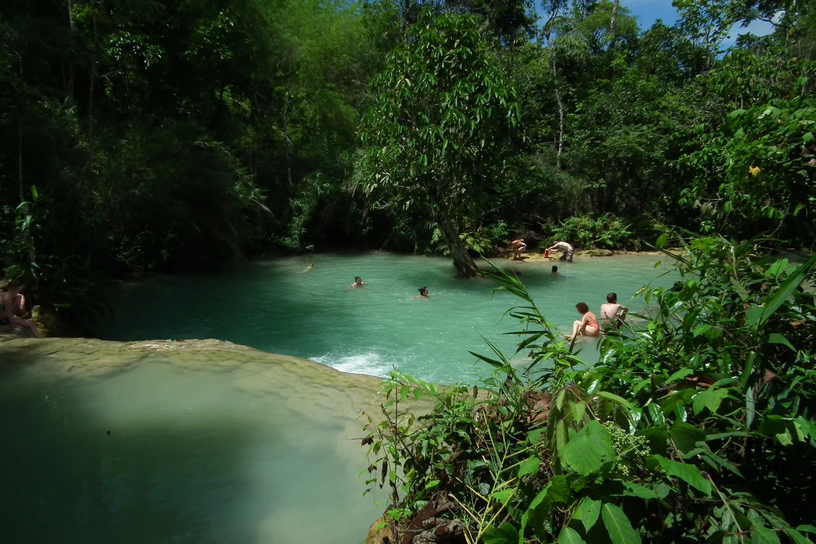 Visitors swim in the turquoise pools of Kuang Si Falls near Luang Prabang, surrounded by lush jungle.