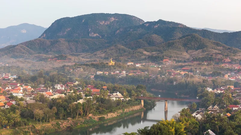 Aerial view over Luang Prabang's rooftops and river bridge framed by forested mountains at dusk.