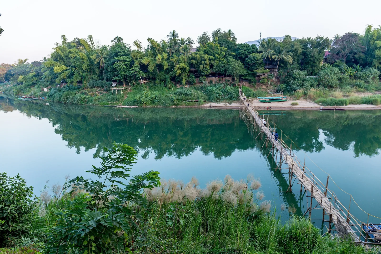 A bamboo footbridge crosses the calm river toward lush greenery near Luang Prabang, Laos.