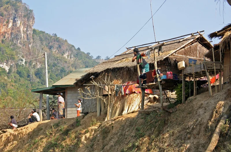 Villagers gather beside thatched-roof stilt houses on a hillside near limestone cliffs in Luang Prabang province