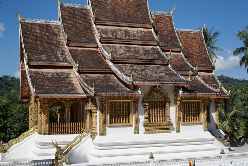A tiered temple roof with golden carvings rises above white walls at a Luang Prabang temple.