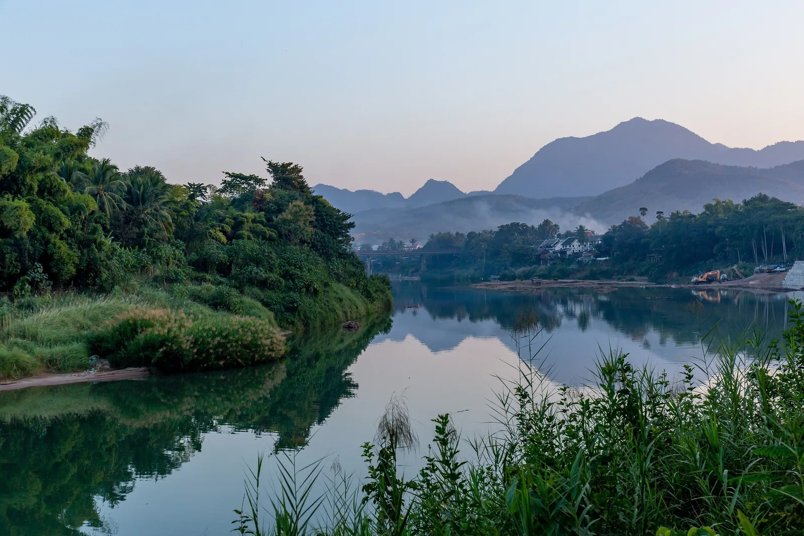 Calm waters of the Nam Khan River reflect green hills and misty mountains near Luang Prabang at dusk. Calm waters of the Nam Khan River reflect green hills and misty mountains near Luang Prabang at dusk.