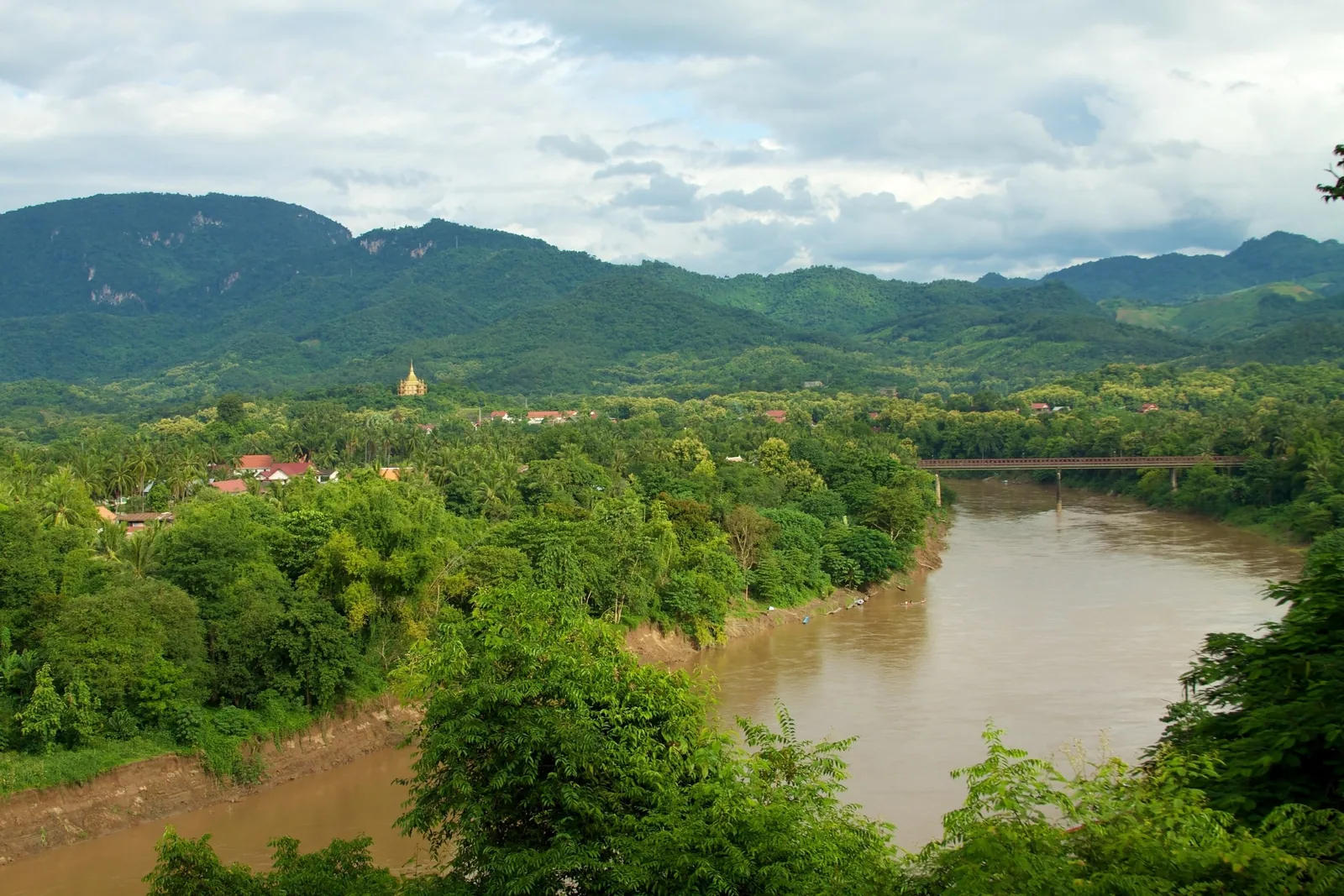 A muddy river winds through green hills past Luang Prabang's rooftops and a distant bridge under cloudy skies.