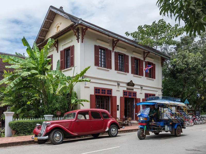 A vintage red car and a tuk-tuk pass a colonial-era building with red shutters in Luang Prabang.