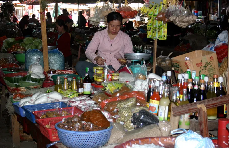 A vendor weighs food at a crowded market stall stocked with bottles, jars and produce in Luang Prabang.