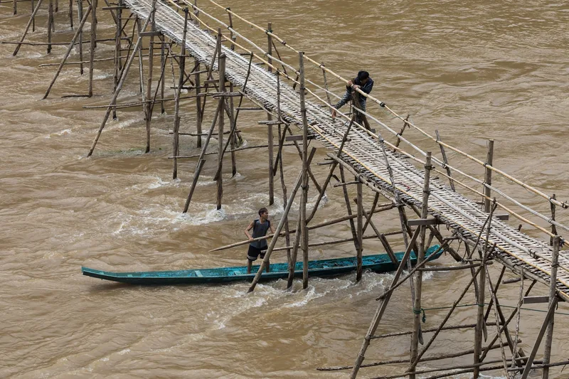 Workers repair a bamboo bridge over the muddy river in Luang Prabang, one steadying a boat below.