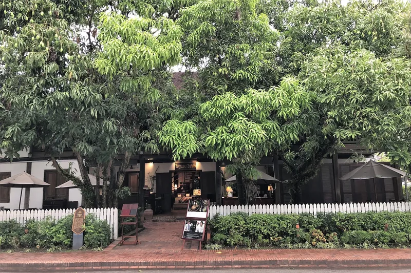 A shaded storefront with dark wooden facade sits behind a white picket fence and large mango trees in Luang Prabang.