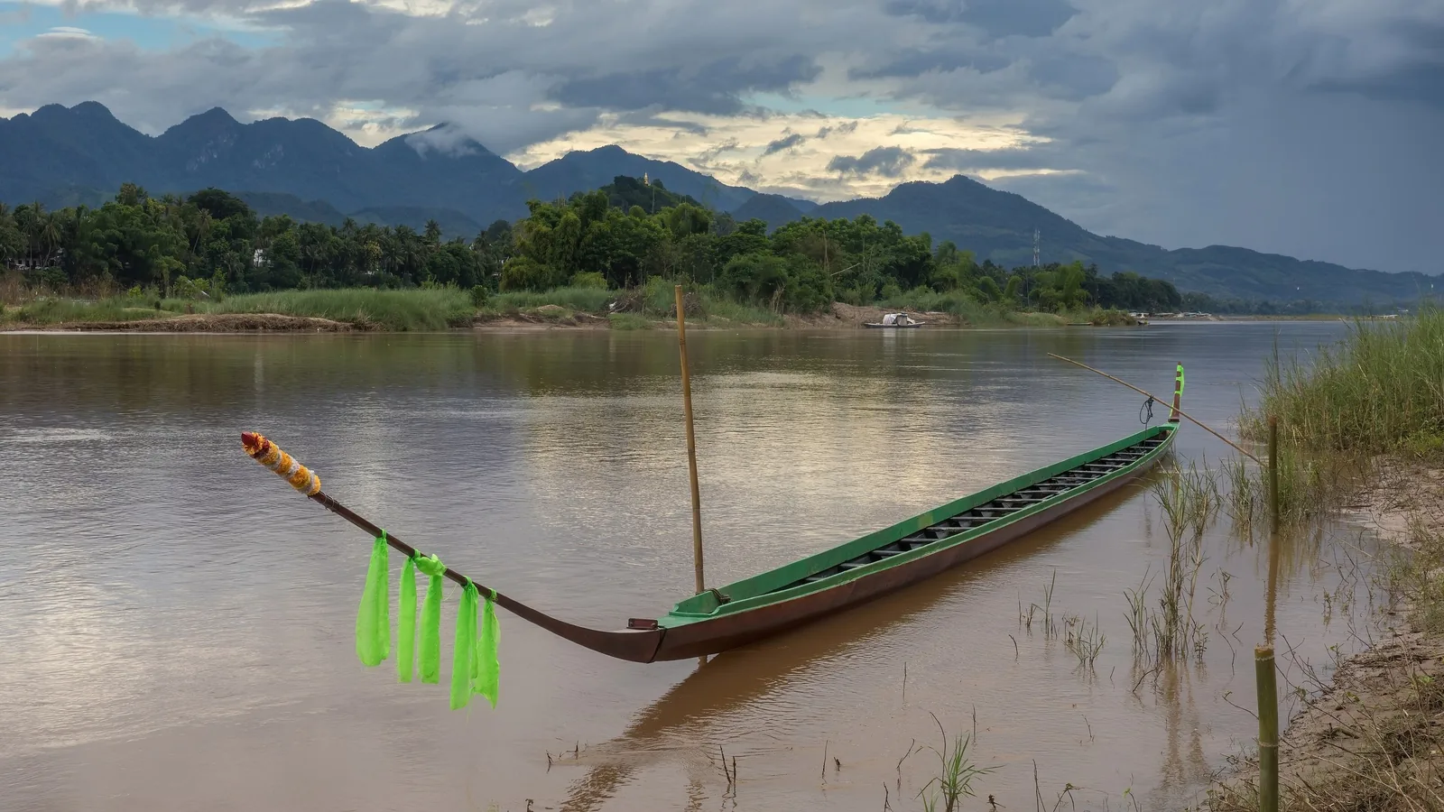 A traditional wooden pirogue with green ribbons rests on the Mekong River near Luang Prabang, mountains rising behind.