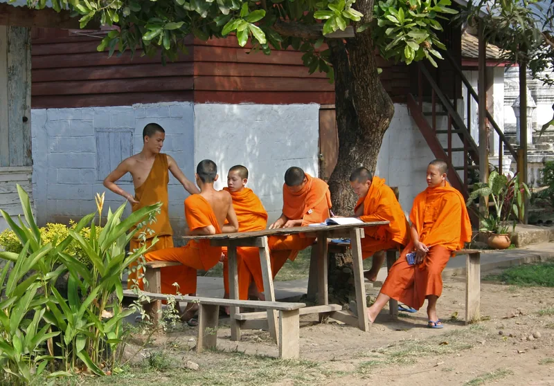 Young Buddhist novice monks in orange robes gather at a wooden table under a tree at a Luang Prabang temple. Young Buddhist novice monks in orange robes gather at a wooden table under a tree at a Luang Prabang temple.