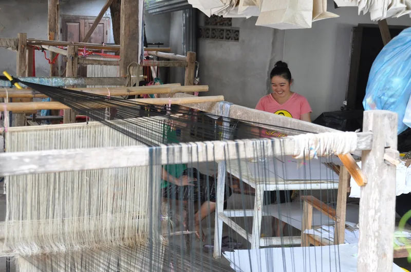 A smiling weaver sits behind a traditional wooden loom strung with threads in a Luang Prabang workshop.