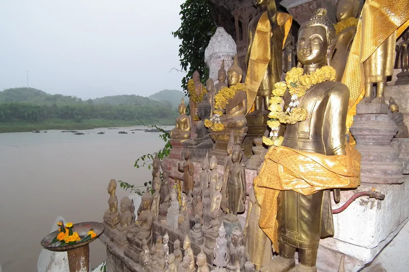 Rows of gilded Buddha statues draped in yellow sashes overlook the Mekong River at Pak Ou Caves near Luang Prabang.