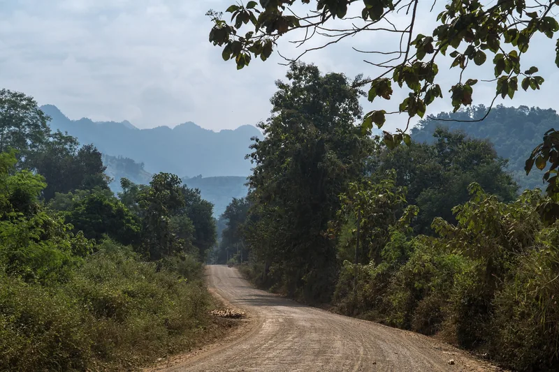 A dirt road winds through dense forest with layered mountains in the background near Luang Prabang.