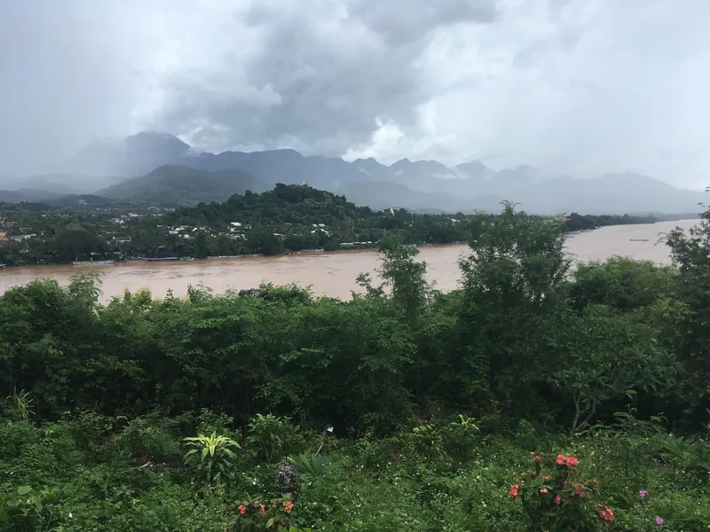 Muddy Mekong River winds past Luang Prabang under misty mountains, seen from a hillside garden.