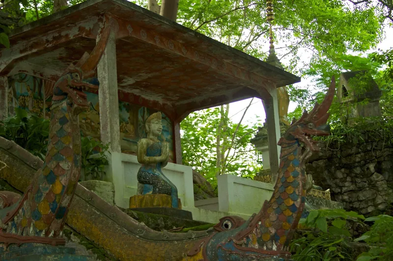 Weathered naga-shaped boat carvings flank a small shrine with a praying figure in Luang Prabang, Laos.