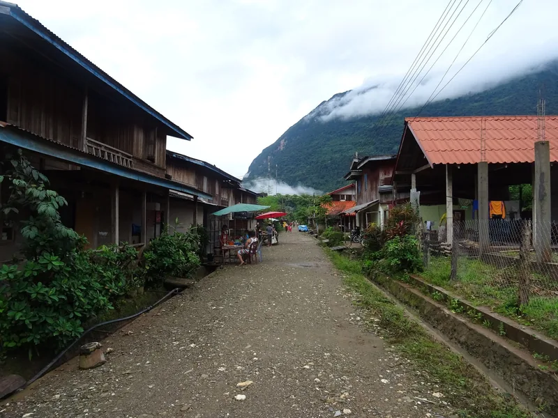 A dirt street lined with wooden houses leads toward misty mountains in Nong Khiaw, Laos.