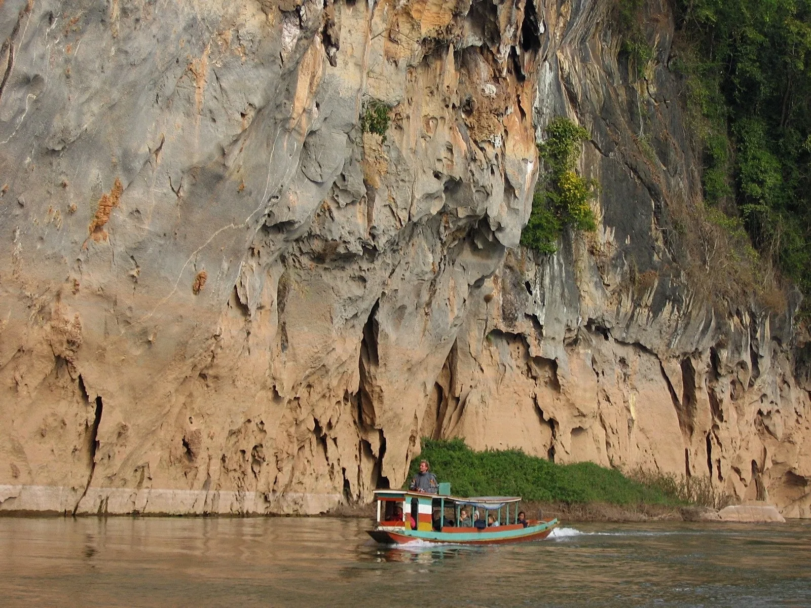 A wooden longtail boat cruises past towering limestone cliffs on the Nam Ou River near Nong Khiaw.