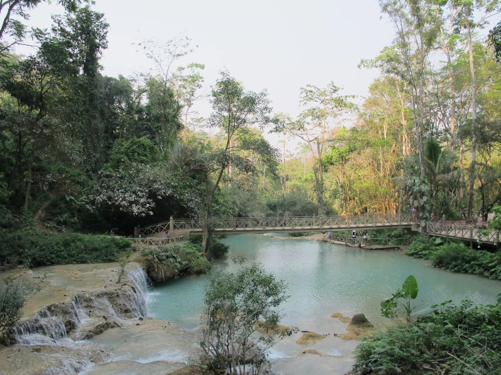 A wooden footbridge crosses turquoise pools at Kuang Si Falls near Luang Prabang, surrounded by lush forest. A wooden footbridge crosses turquoise pools at Kuang Si Falls near Luang Prabang, surrounded by lush forest.