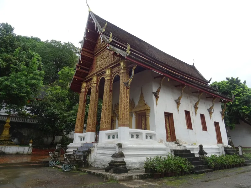 A white temple hall with gilded pillars and a multi-tiered roof stands among trees in Luang Prabang, Laos.