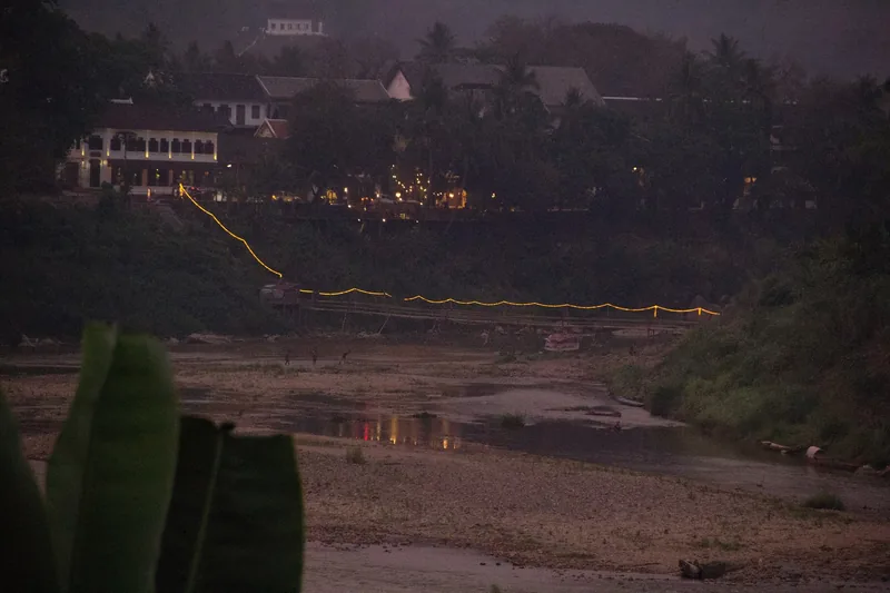 A string of lights traces a bamboo footbridge over a dry riverbed at dusk in Luang Prabang.