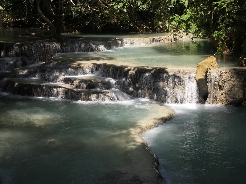 Tiered turquoise pools cascade over limestone shelves at Kuang Si Waterfall near Luang Prabang, surrounded by dense forest.