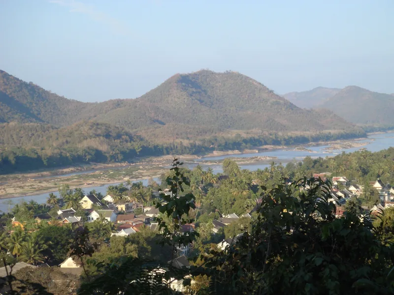 A hillside view over Luang Prabang's rooftops toward the Mekong River and forested mountains beyond.
