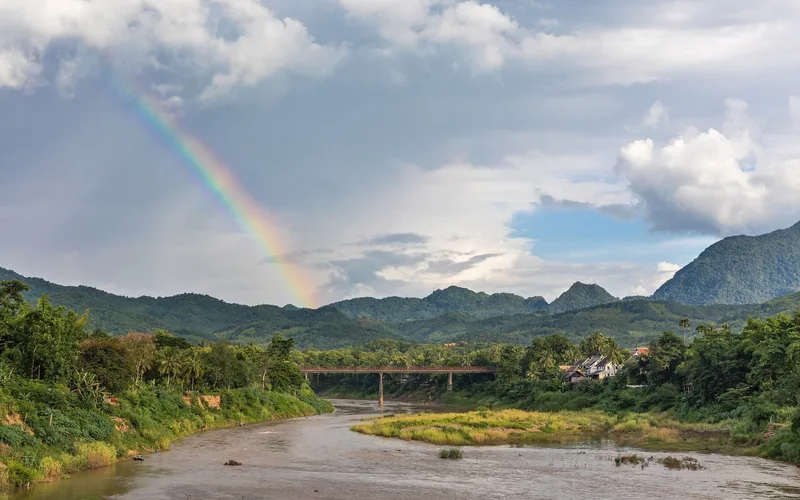 A rainbow arcs over a river, old bridge, and forested mountains near Luang Prabang, Laos.