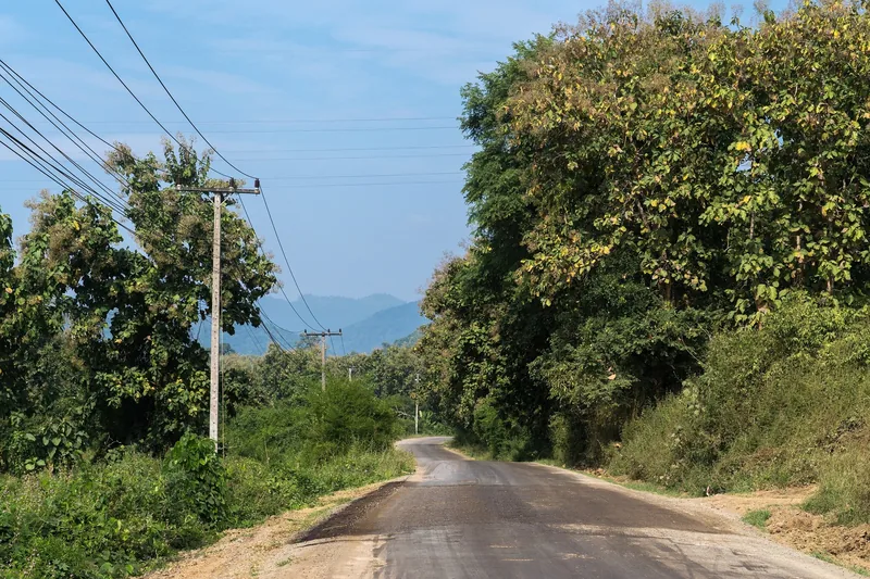 A quiet rural road winds through forested hills near Luang Prabang, with power lines running alongside.