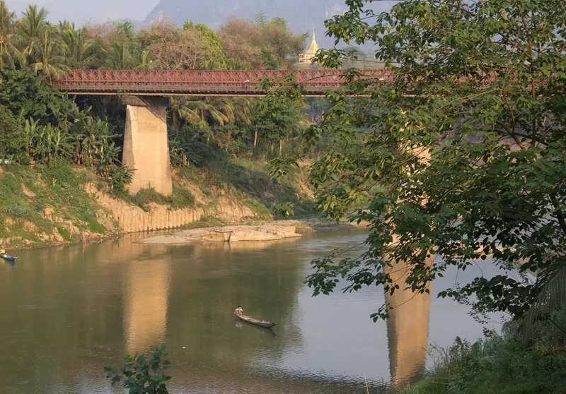 A red steel bridge spans the Nam Khan River near Luang Prabang, with a small boat drifting below.