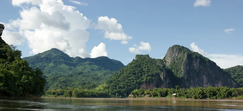 Green mountains rise above the Mekong River near Luang Prabang under a partly cloudy sky.