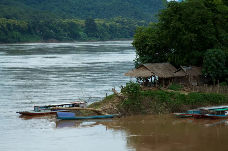 Wooden boats moored along the muddy Mekong riverbank near a thatched hut in Luang Prabang, Laos.