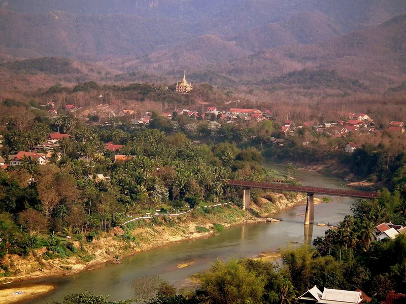 An elevated view over Luang Prabang shows a river bridge, golden temple spire, and rooftops amid green hills.