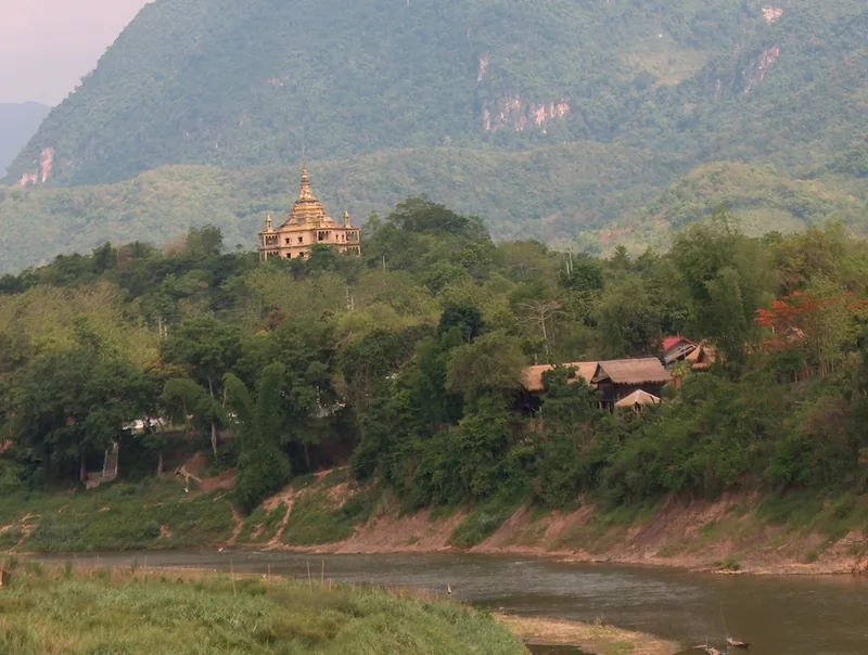 A golden temple spire rises above riverside trees and hills along the Khan River near Luang Prabang.