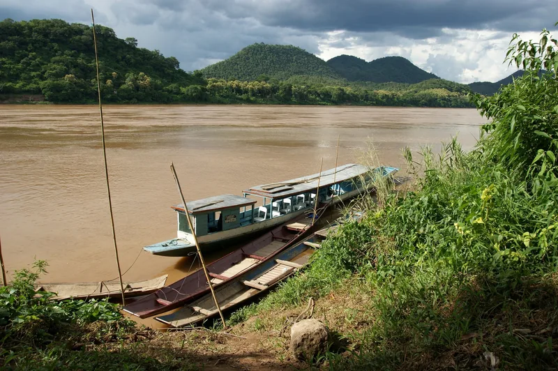 Long-tail boats moored on the muddy Mekong River banks near Luang Prabang, with forested hills in the background. Long-tail boats moored on the muddy Mekong River banks near Luang Prabang, with forested hills in the background.