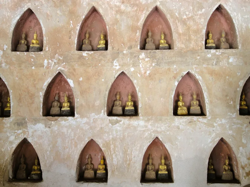 Rows of small gilded Buddha statues rest in arched niches on a weathered temple wall in Vientiane, Laos. Rows of small gilded Buddha statues rest in arched niches on a weathered temple wall in Vientiane, Laos.