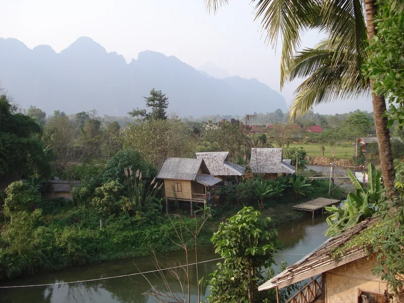 Wooden stilt houses beside a river with limestone karst mountains rising in the misty background near Vang Vieng.