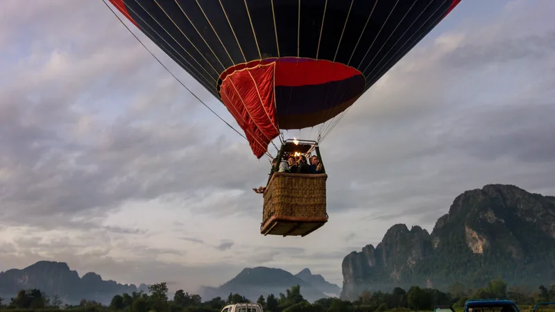 A hot air balloon with a basket of passengers rises over the limestone karsts of Vang Vieng at dawn.